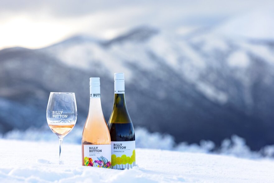 Two glasses of wine and a wine glass nestled on a snowy hilltop with snowtopped mountains in the background.