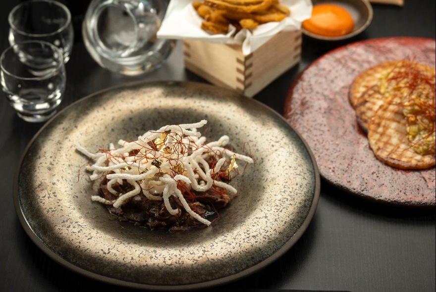 A Japanese dish with crispy noodles, on a traditional Japanese plate on a black table, with other plates, dishes and glassware surrounding it.