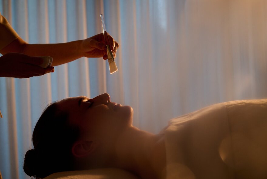 The silhouette of a person enjoying a spa body treatment at the Bamford Wellness Spa, a hand hovering over their face. 