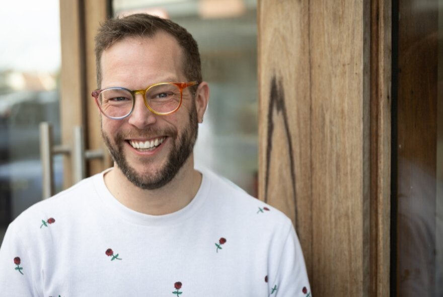 Bearded man with pink glasses, smiling, wearing a white top with floral detail.