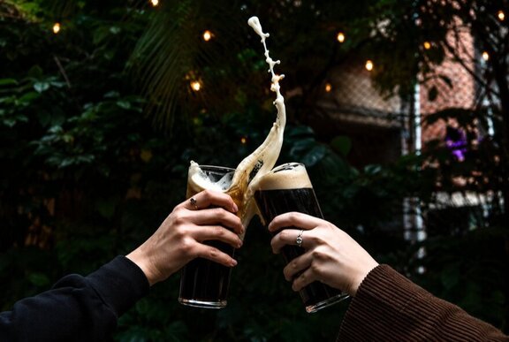 Two hands bring two pints of dark stout beer together in a clinking gesture, with foam from one spilling out.