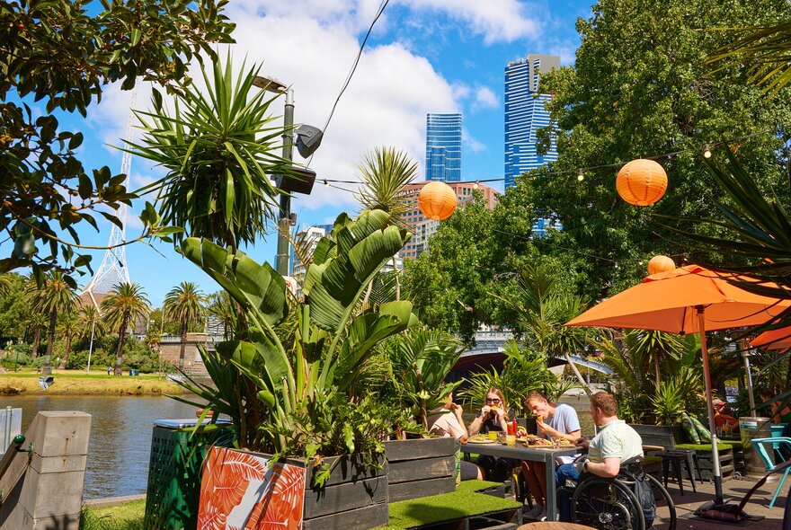 A group of friends are eating lunch at an outdoor bar by the river. There are two people in a wheelchair. 