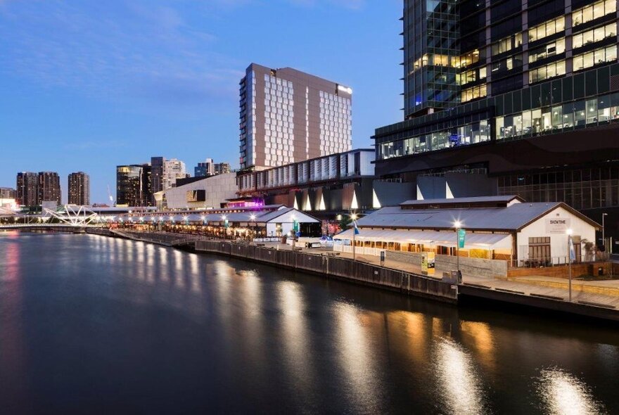 Cargo sheds on the river with tall buildings in the background, lit up at dusk.