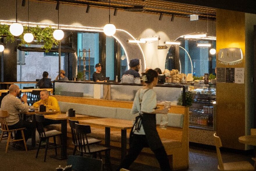 Interior of a cafe with people seated at a table eating and drinking, a waiter walking around and a service counter in the background. 