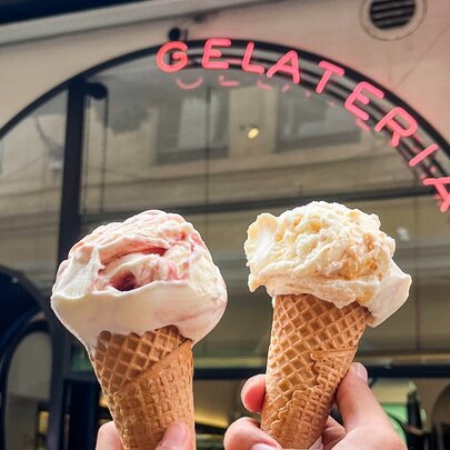 Hands holding gelati cones in front of a Gelateria sign.