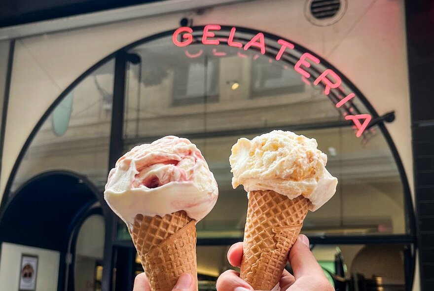 Hands holding gelati cones in front of a Gelateria sign.