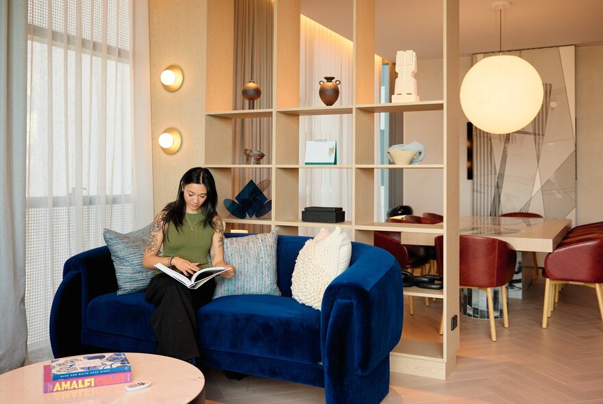 A person is sitting in a modern hotel room reading a book on a blue velvet sofa. Behind them is a shelf with decor items. 