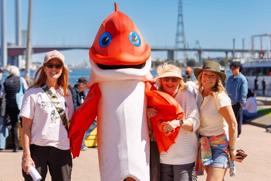 People posing with a red fish mascot at a community or waterfront event such as a farmer's market.
