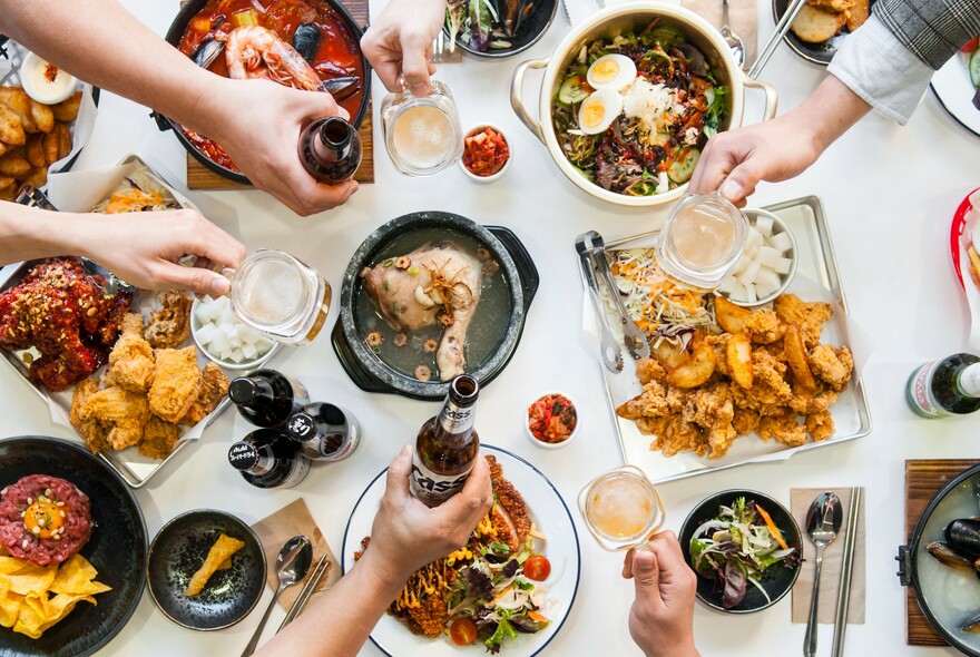 Dishes of chicken and sides on white table with hands clinking glasses and bottles.