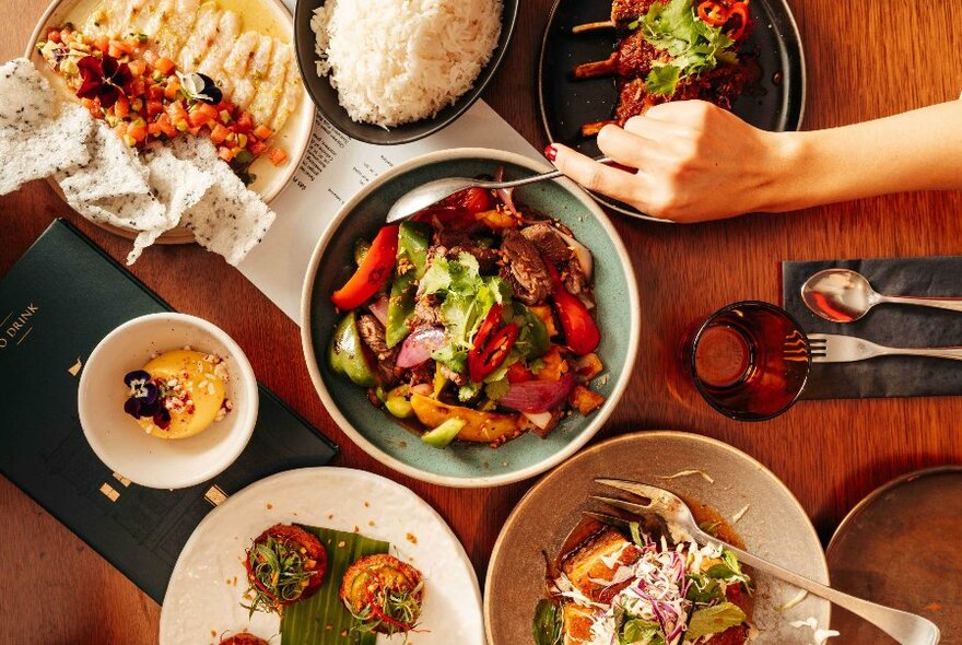 Several plates of food on a table, with glasses and cutlery, with a hand holding a spoon reaching into a bowl of salad, seen from above.