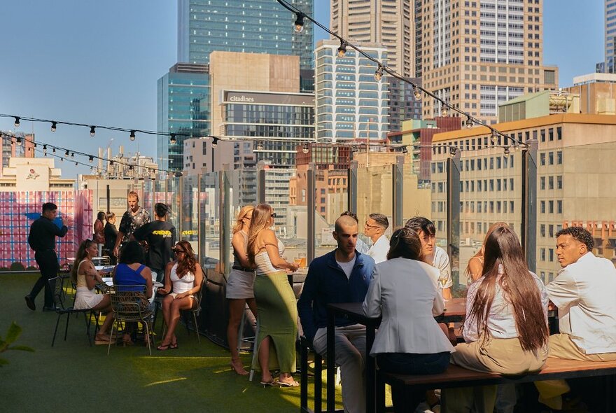 People socialising and drinking in the outdoor bar at Blossom rooftop, with a view of city buildings in the background.