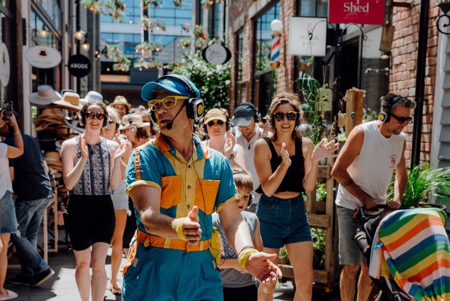 A musical walking tour being led through a Melbourne laneway by a man wearing a blue cap, headphones, speaker and overalls.