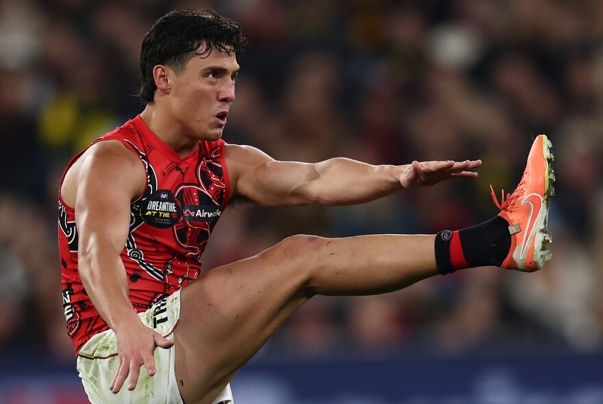 An Essendon AFL football player with his leg raised high in the air, having just kicked the footy.