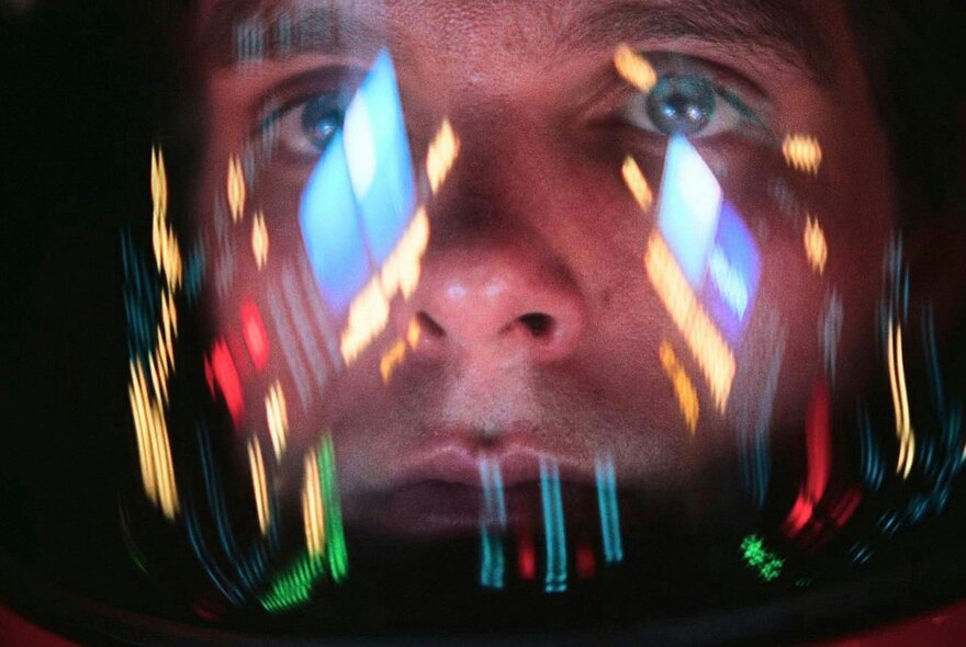 A man's face seen behind a clear helmet with neon lights reflected in it.