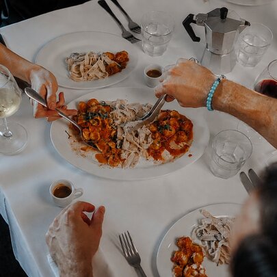 Two diners at a white cloth-covered table, helping themselves to Pasta Mista, a plate of three different types of pasta, with wine glasses and espresso cups on the table.