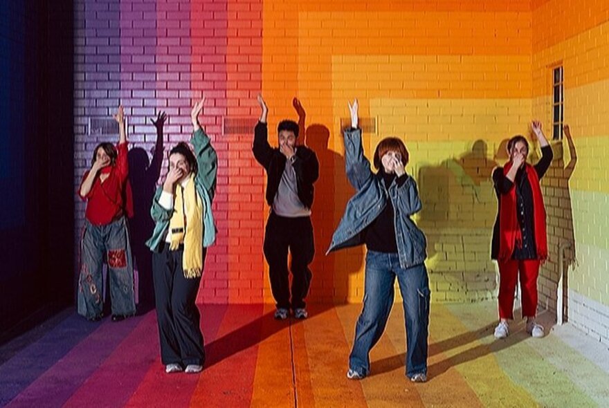 Five dancers each doing the snorkel dance-move, standing on a rainbow-coloured floor in front of a rainbow-coloured painted wall.