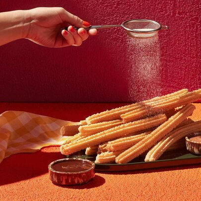 A hand sprinkling icing sugar over a platter of churros.