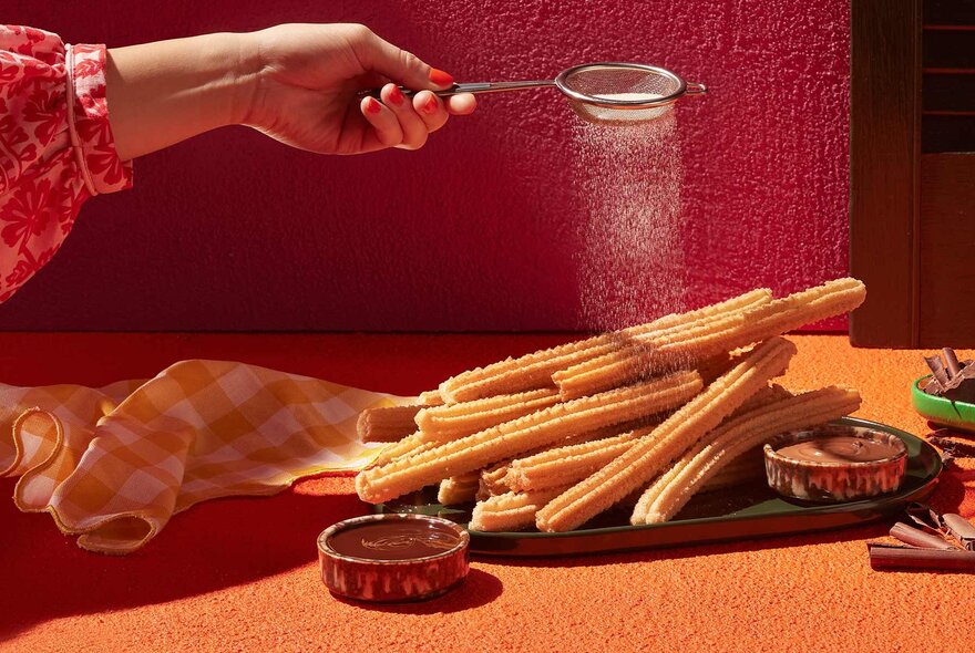 A hand sprinkling icing sugar over a platter of churros.