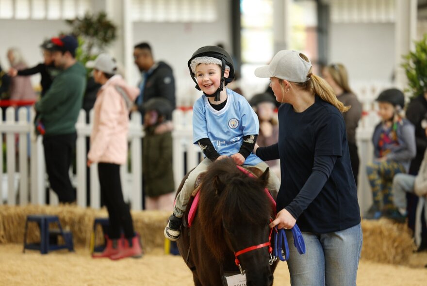 A smiling young boy sitting on a pony being led around an indoor enclosure by a pony handler, people lined up in the background.