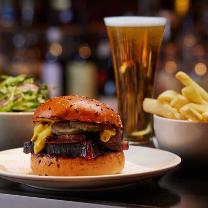 A 12-hour smoked beef brisket burger, bowl of chips, salad and a pot of beer placed at a bar.