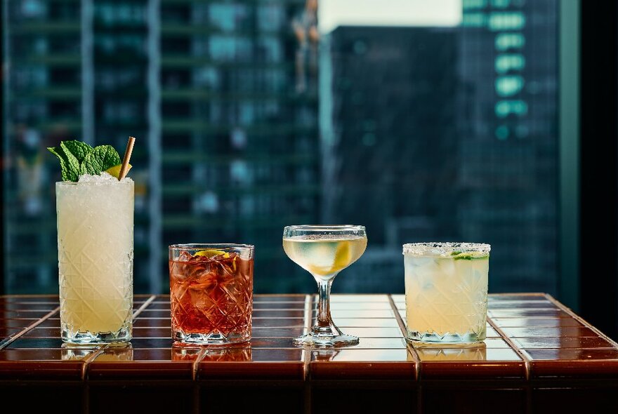 Four different cocktails presented on a tiled surface, in front of a window with Melbourne city buildings in the background.