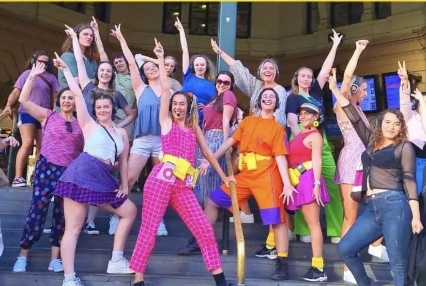 Group of people wearing headphones and posing with one arm raised in the air above their heads in a dance-style move, on the steps of Flinders Street station.