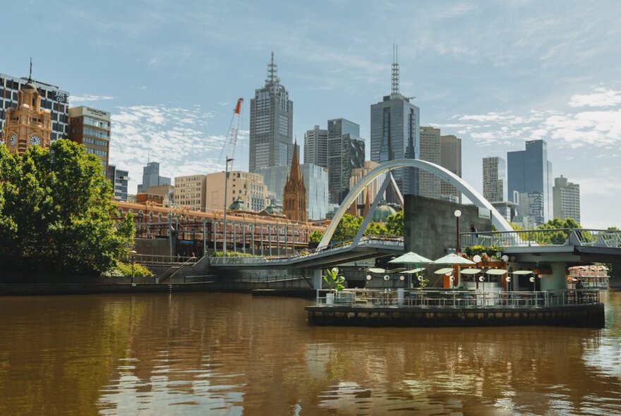 The floating bar Ponyfish Island in the middle of the Yarra River with a bridge and a view of the city's building skyline in the background.