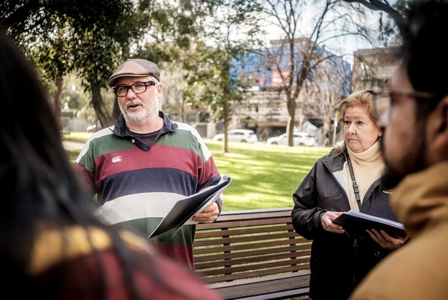 A tour guide leading a walking street tour, talking outdoors in front of a small nature reserve with a few people listening.