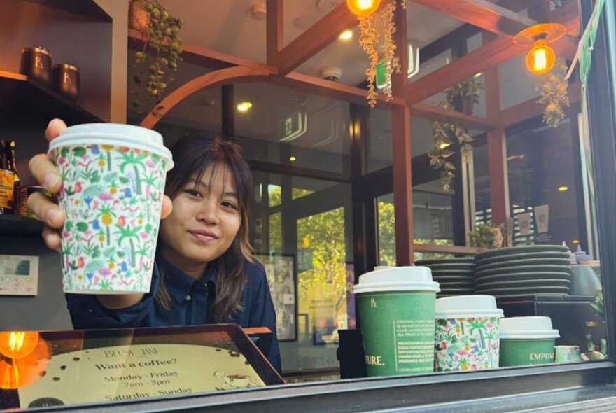 A woman in a small cafe window holding forth a takeaway coffee cup for someone to take. 