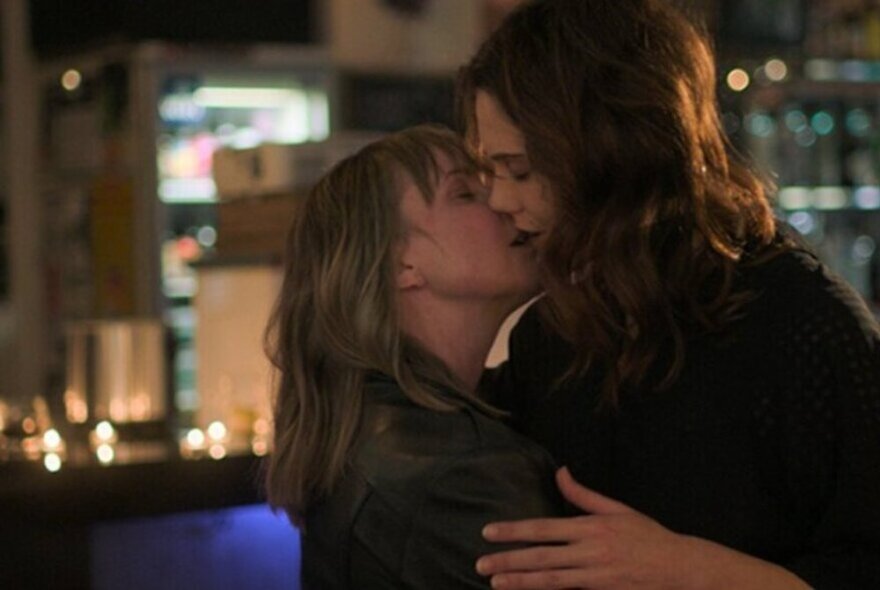 Movie still, female couple embracing in a kiss in a candle-lit room with city lights in the background.