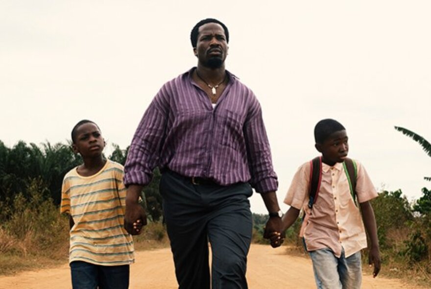 A still from an African film showing a father and his two sons walking down a dirt road holding hands.