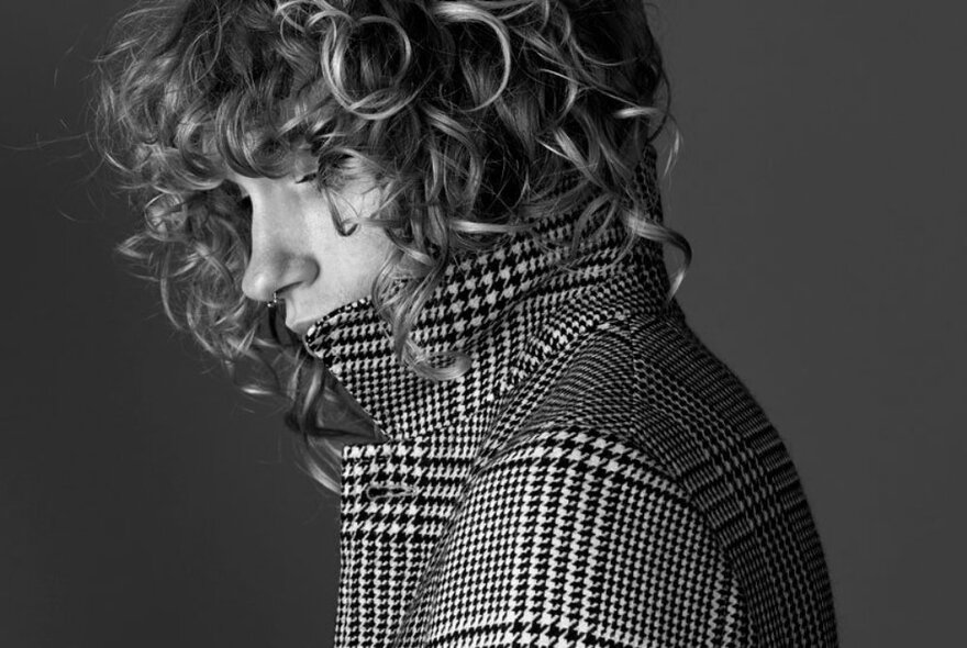 Handsome young male model, with curled golden hair, in side profile, wearing a black and white houndstooth heavy overcoat.