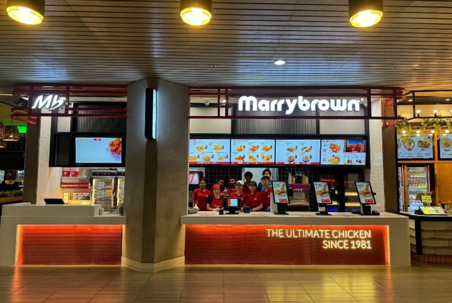 The front of the Marrybrown open food kiosk in Melbourne Central's food court, showing a service counter, a large illuminated menu on the rear wall and service personnel.