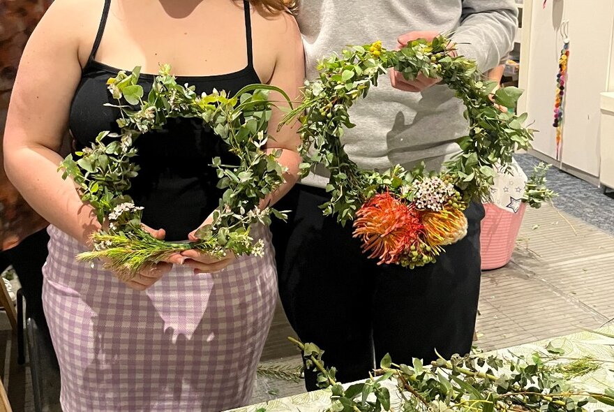 Two people standing in front of a work table, holding up circular wreaths they've made using native flowers and greenery.