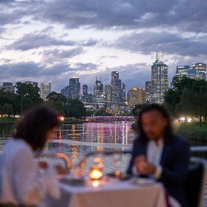 A blurred image of two people at a private table with a candle on the river with the city in the background. 