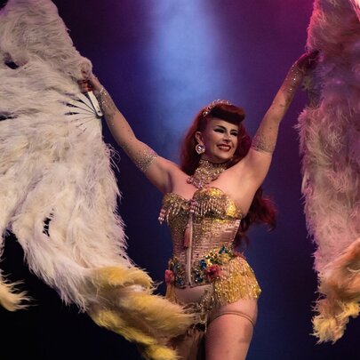 A burlesque dancer in a corset holding up two large feathered.