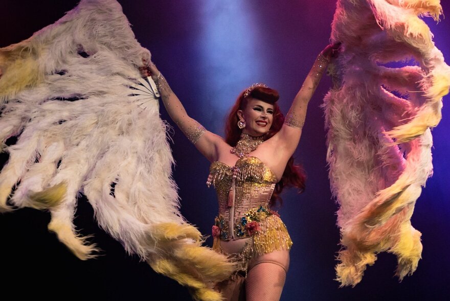 A burlesque dancer in a corset holding up two large feathered.