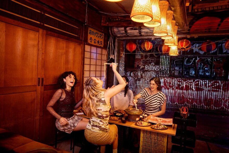 People seated at a table in a private booth in a Japanese restaurant with sliding doors and lights, one person draping noodles into her mouth.