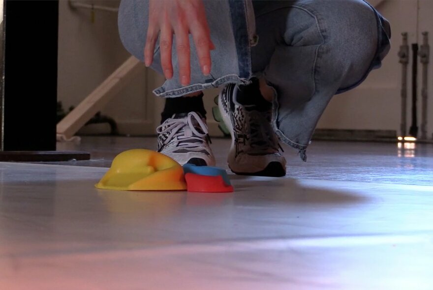 A figure squats over three rubber mounds on a white floor, the figure is only visible from ankles down, in blue jeans and white and black running shoes.