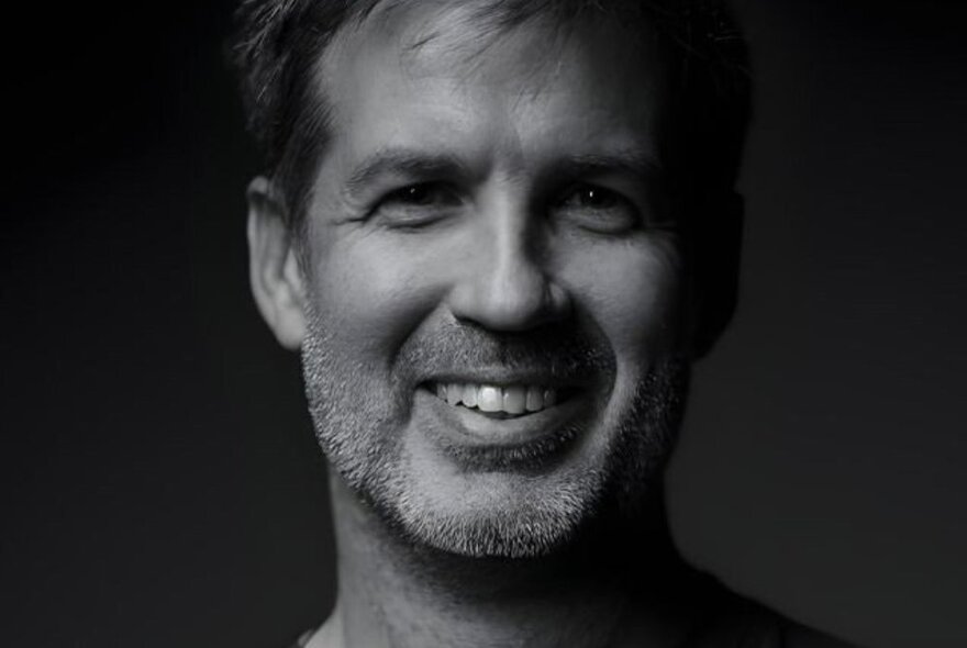 Man with stubbled face smiling against dark background.