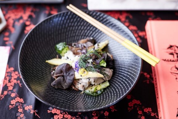 A dish of food served in a shallow black bowl, with chopsticks resting on the side of the bowl.
