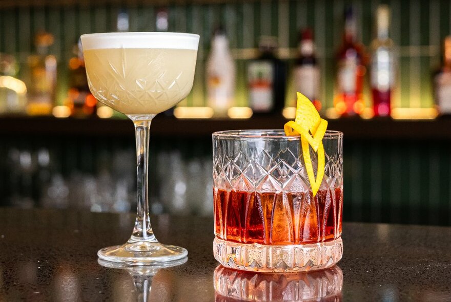 One tall cocktail in a cut crystal wine glass and a short cocktail in a cut crystal tumbler with a lemon garnish.