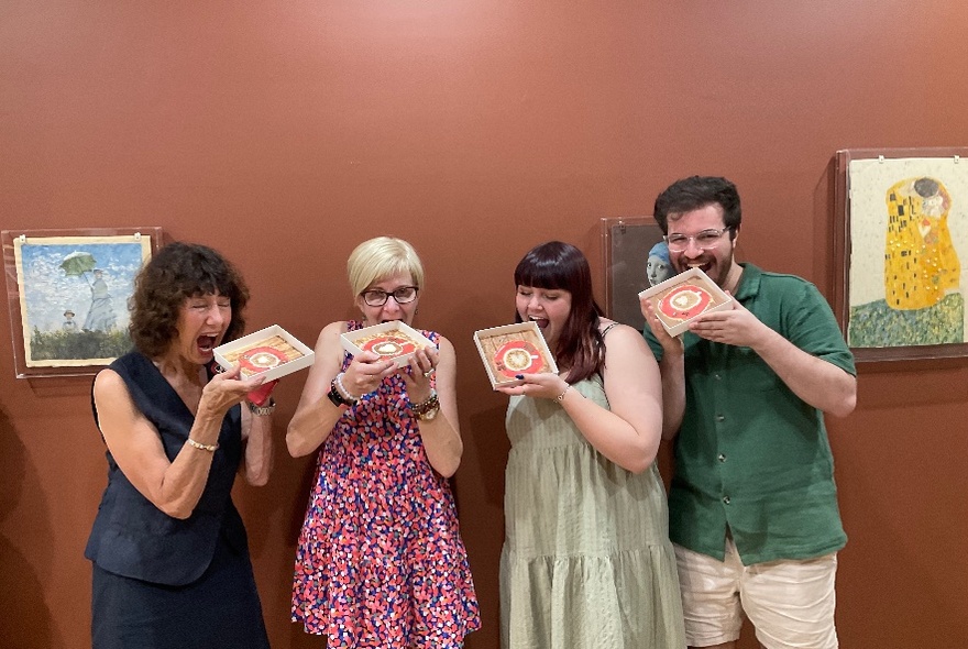 A row of people holding up painted squares, about to take a bite, in front of a wall of canvasses.