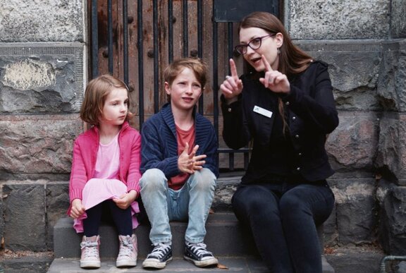 Adult seated with two children on a stone step, making an outline with her fingers.