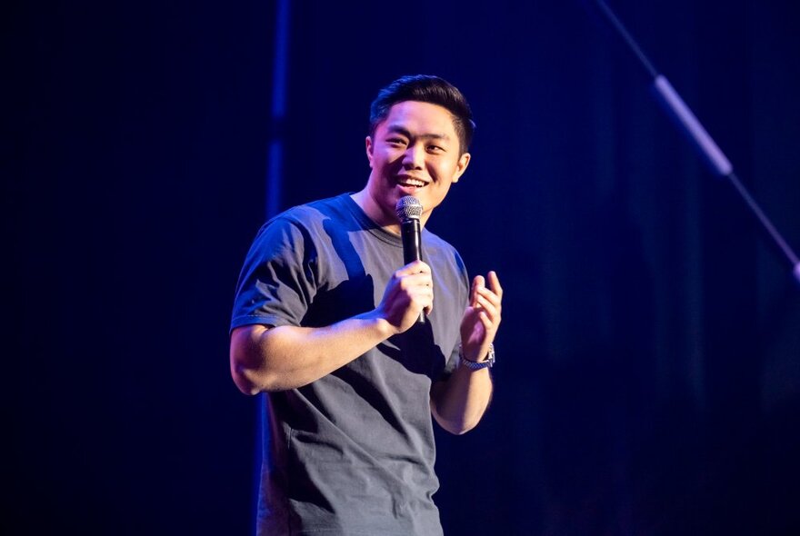 A smiling young man standing on a stage, talking into a microphone he holds in his hand.