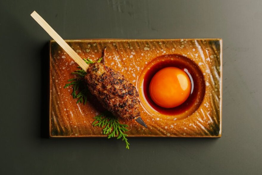 A single Japanese yakitori skewer on a rectangle platter with a circular depression holding a raw egg yolk.