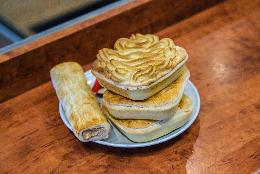 Three pies and a sausage roll on a white plate on a wooden counter inside a cafe.