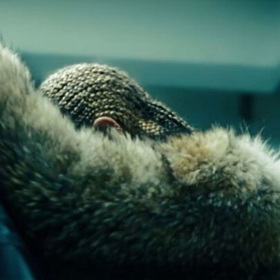 A still from Beyoncé's visual album Lemonade, depicting the singer in profile leaning against a car, wearing a fur jacket, her hair braided, her face covered.