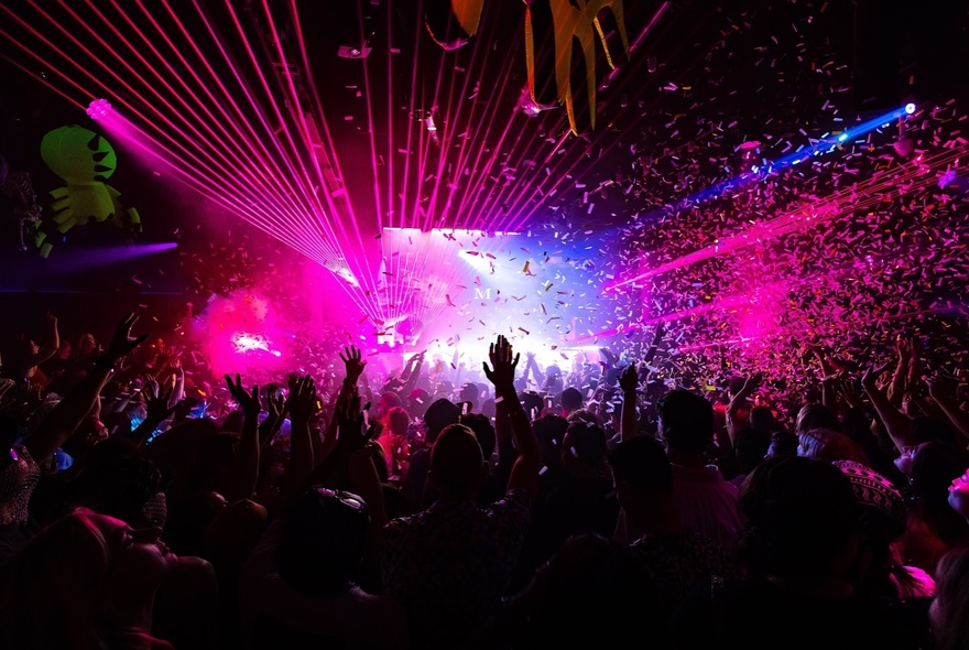 A dance club setting with a crowd enjoying a DJ set and pink lights on the stage. 