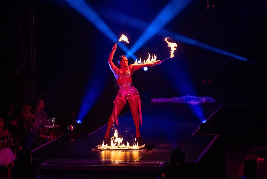A fire performer in a glittering red outfit dances on a dark cabaret stage, surrounded by flames on the floor and holding two flaming torches, illuminated by bright blue spotlights. 